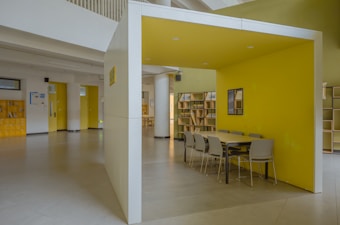A modern indoor space featuring a yellow-walled booth with a long table and several gray chairs. The area is well-lit and spacious, with minimalistic design elements. Bookshelves line the walls, and there are various doors leading to adjacent rooms.
