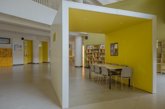 A modern indoor space featuring a yellow-walled booth with a long table and several gray chairs. The area is well-lit and spacious, with minimalistic design elements. Bookshelves line the walls, and there are various doors leading to adjacent rooms.