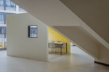 A minimalist indoor space featuring a geometric design with clean lines. A rectangular structure with a small window is positioned under a large staircase. The structure has a bright yellow wall visible through an open side, with a table and chairs set up inside. The floor is tiled in a light color, and large windows on an adjacent building can be seen in the background.