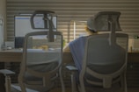 A person is seated at a desk, viewed from behind through two mesh office chairs. The workspace features a computer monitor and a laptop, with soft ambient lighting from a desk lamp creating a warm atmosphere. The room is lined with horizontal blinds, indicating a modern office setup.