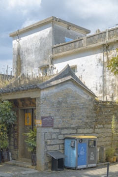 A rustic building with weathered white walls and a traditional style roof. Vegetation is visible on the roof, while a wooden door is framed by potted plants. There are blue waste and recycling bins near the entrance, indicating environmental awareness. The structure seems to have historical or cultural significance.