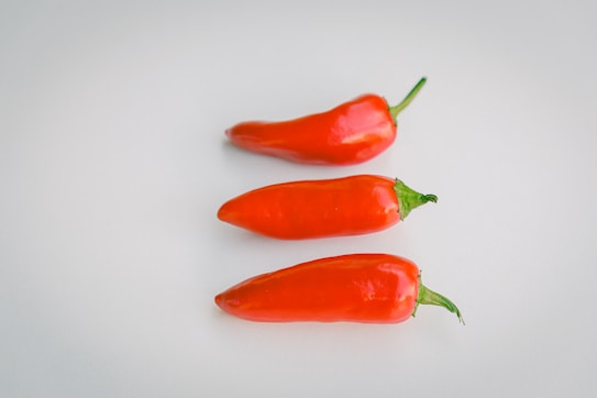 Three vibrant red chili peppers arranged neatly in a vertical line on a smooth, light-colored surface. Each chili pepper has a glossy texture and a fresh green stem.