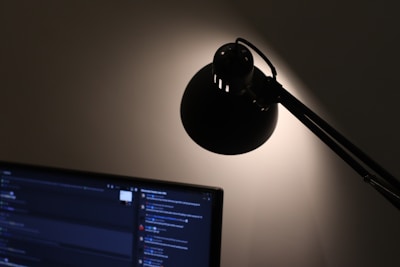 A dark room with a computer monitor displaying a dark-themed interface on the left side and a desk lamp casting light on the wall.