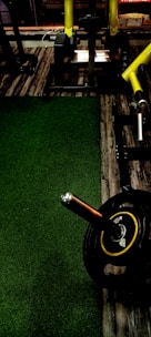 A gym setting with exercise equipment, including a barbell with weights on a stand. The floor features a combination of wood and green carpet. There are yellow handles on the equipment and an industrial aesthetic is present.