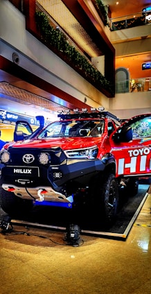 A vibrant red Toyota Hilux pickup truck is displayed in a well-lit indoor setting, possibly a showroom or a mall. The vehicle is equipped with off-road enhancements like a winch, large tires, and roof-mounted lights. The background shows multiple floors with railings and greenery.
