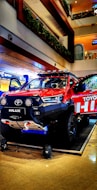 A vibrant red Toyota Hilux pickup truck is displayed in a well-lit indoor setting, possibly a showroom or a mall. The vehicle is equipped with off-road enhancements like a winch, large tires, and roof-mounted lights. The background shows multiple floors with railings and greenery.