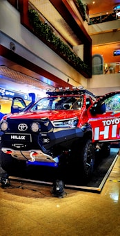 A vibrant red Toyota Hilux pickup truck is displayed in a well-lit indoor setting, possibly a showroom or a mall. The vehicle is equipped with off-road enhancements like a winch, large tires, and roof-mounted lights. The background shows multiple floors with railings and greenery.