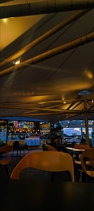 A dimly lit fast-food restaurant with a modern design featuring exposed ducts and pipes on the ceiling. There are empty yellow chairs and tables in the foreground. The counter in the background is illuminated with menu boards displaying colorful options. There are a few customers visible near the counter and in the outdoor seating area.