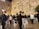 Groups of people are gathered at the Western Wall, dressed in traditional black attire and hats. Some are standing and others are seated on white plastic chairs, engaged in prayer or reflection. The ancient stone wall is illuminated by soft lighting, with small patches of greenery growing between the stones.