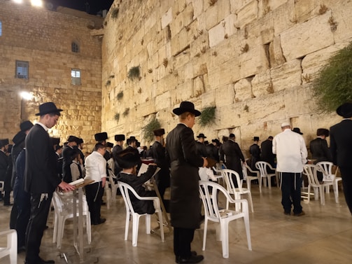 Groups of people are gathered at the Western Wall, dressed in traditional black attire and hats. Some are standing and others are seated on white plastic chairs, engaged in prayer or reflection. The ancient stone wall is illuminated by soft lighting, with small patches of greenery growing between the stones.