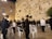 Groups of people are gathered at the Western Wall, dressed in traditional black attire and hats. Some are standing and others are seated on white plastic chairs, engaged in prayer or reflection. The ancient stone wall is illuminated by soft lighting, with small patches of greenery growing between the stones.