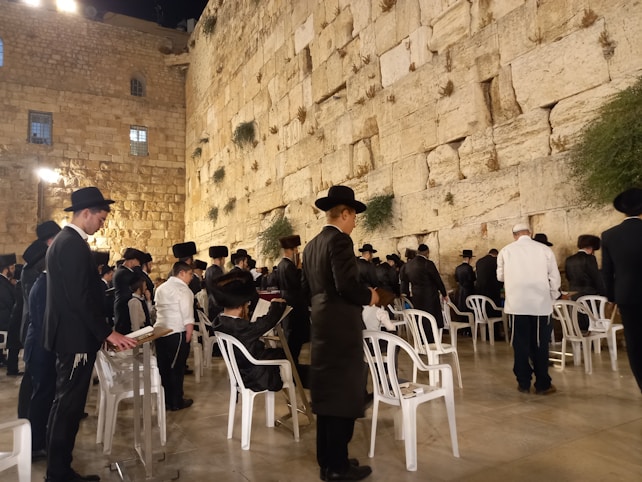 Groups of people are gathered at the Western Wall, dressed in traditional black attire and hats. Some are standing and others are seated on white plastic chairs, engaged in prayer or reflection. The ancient stone wall is illuminated by soft lighting, with small patches of greenery growing between the stones.