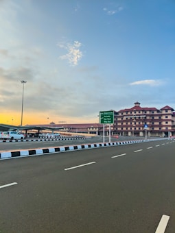 A wide road in front of an airport building with a clear sky at sunset. Several cars are parked on the left side of the road. The airport building has a distinct architectural style with multiple floors and a reddish-brown color. A large green sign gives directions for departures and to the car park.