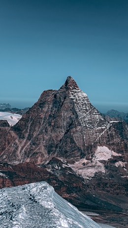 A majestic, snow-capped mountain peak rises prominently against a clear blue sky. The rugged texture of the cliffs contrasts with the smooth, white snow that highlights the contours of the mountain. The surrounding landscape includes additional peaks and valleys, creating a vast and expansive feeling.