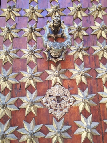 An ornate door with intricate metalwork, featuring several gold-colored star-shaped studs arranged symmetrically across a rich wooden background. The centerpiece is a bronze or brass door knocker in the shape of a mythical creature surrounded by detailed designs, complementing a decorative keyhole cover below.