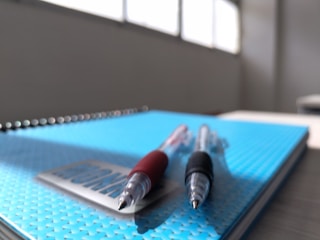 Two ballpoint pens, one with a red grip and the other with a black grip, lay on top of a closed blue notebook on a desk. The background is softly blurred, highlighting a set of windows allowing natural light into the room.