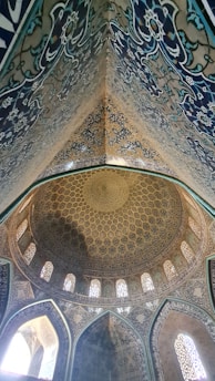 A highly ornate ceiling with intricate geometric patterns and detailed floral designs, featuring a large, domed structure with arched windows allowing natural light to illuminate the rich blues, golds, and whites of the decor. The symmetry and complexity of the artwork reflect traditional Islamic architectural influences.