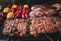 A close-up of a grill loaded with various meats and vegetables. On the left, several whole bell peppers in red and yellow are placed. The center and right are occupied by different cuts of meat, including sausages and steaks. The grill marks are visible on the meats and vegetables as they cook over the heat.