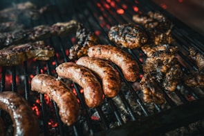 Various meats, including sausages and grilled patties, are cooking on a grill over glowing coals. The grill marks are visible on the browned surfaces, and the embers beneath cast a warm glow.