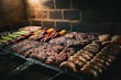 An assortment of meats and vegetables is being grilled on a barbecue. The variety includes sausages, steaks, chicken, and colorful vegetables like bell peppers and zucchini, neatly arranged over a metal grill surface. The background features a rustic brick wall, and the lighting suggests a warm and inviting atmosphere.