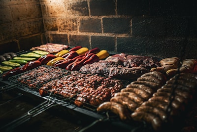 An assortment of meats and vegetables is being grilled on a barbecue. The variety includes sausages, steaks, chicken, and colorful vegetables like bell peppers and zucchini, neatly arranged over a metal grill surface. The background features a rustic brick wall, and the lighting suggests a warm and inviting atmosphere.