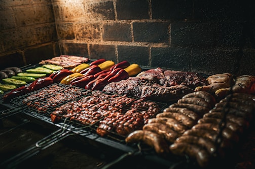 An assortment of meats and vegetables is being grilled on a barbecue. The variety includes sausages, steaks, chicken, and colorful vegetables like bell peppers and zucchini, neatly arranged over a metal grill surface. The background features a rustic brick wall, and the lighting suggests a warm and inviting atmosphere.