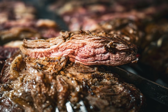 A close-up view of a juicy, grilled steak with visible grill marks and a seared exterior. The meat appears tender and succulent with a well-defined texture. A sharp knife can be seen slicing through the steak, highlighting its pink interior.