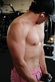 A shirtless man with earphones is wearing a black cap while lifting weights. His muscular physique is prominent, and he is positioned in what appears to be a gym setting. The focus is on his upper body and arms, which display defined muscles. He is looking downwards, possibly concentrating on the exercise.