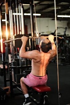 A person is engaged in a workout, performing pull-ups on a resistance machine in a gym setting. The individual is wearing pink shorts and a cap, facing away from the camera with a visible muscular back. The gym is equipped with various exercise machines, and warm lighting creates a focused ambiance.
