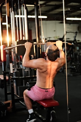 A person is engaged in a workout, performing pull-ups on a resistance machine in a gym setting. The individual is wearing pink shorts and a cap, facing away from the camera with a visible muscular back. The gym is equipped with various exercise machines, and warm lighting creates a focused ambiance.