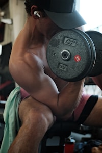 A person is engaged in a bicep curl exercise, holding a heavy dumbbell in one hand. They are wearing a cap, earphones, and shorts, seated on a bench in an indoor gym setting. The focus is on their muscular arm and the 35-pound weight.