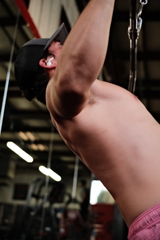 A person wearing a cap and earphones is performing a pull-up exercise in a gym setting. The focus is on their upper body and arm muscles, with gym equipment and fluorescent lights visible in the background.