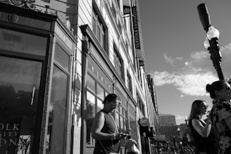 A street scene with several people standing on the sidewalk next to a multi-story building. The building has large windows and ornate architectural details. Signs indicate that the building is affiliated with Suffolk University. A tall street lamp is visible, casting shadows on the building. The sky is clear with some clouds.