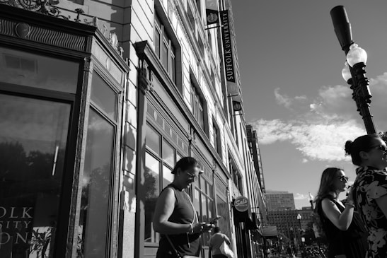 A street scene with several people standing on the sidewalk next to a multi-story building. The building has large windows and ornate architectural details. Signs indicate that the building is affiliated with Suffolk University. A tall street lamp is visible, casting shadows on the building. The sky is clear with some clouds.