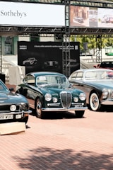 A collection of classic cars is displayed outdoors on a brick-paved area. Behind the cars, there is a banner with 'Sotheby's' and other automotive images. The cars, polished and well-maintained, include a green car in the center with a distinctive front grill, flanked by other vintage vehicles.