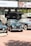 A collection of classic cars is displayed outdoors on a brick-paved area. Behind the cars, there is a banner with 'Sotheby's' and other automotive images. The cars, polished and well-maintained, include a green car in the center with a distinctive front grill, flanked by other vintage vehicles.