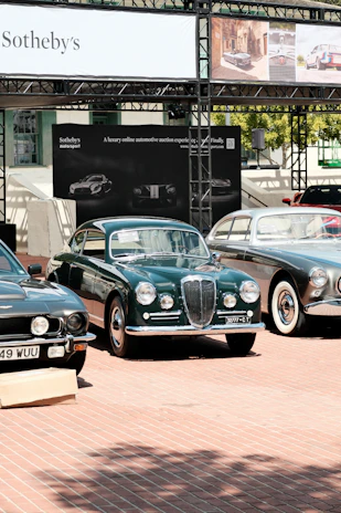 A collection of classic cars is displayed outdoors on a brick-paved area. Behind the cars, there is a banner with 'Sotheby's' and other automotive images. The cars, polished and well-maintained, include a green car in the center with a distinctive front grill, flanked by other vintage vehicles.