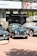 A collection of classic cars is displayed outdoors on a brick-paved area. Behind the cars, there is a banner with 'Sotheby's' and other automotive images. The cars, polished and well-maintained, include a green car in the center with a distinctive front grill, flanked by other vintage vehicles.