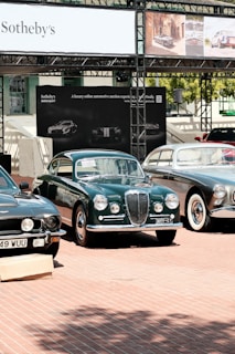 A collection of classic cars is displayed outdoors on a brick-paved area. Behind the cars, there is a banner with 'Sotheby's' and other automotive images. The cars, polished and well-maintained, include a green car in the center with a distinctive front grill, flanked by other vintage vehicles.
