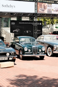 A selection of classic cars displayed outdoors on a brick platform, beneath a large Sotheby's auction sign. The scene includes a row of well-maintained vintage vehicles, including green and black models, with a background of promotional banners for a luxury online automotive auction.