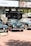 A selection of classic cars displayed outdoors on a brick platform, beneath a large Sotheby's auction sign. The scene includes a row of well-maintained vintage vehicles, including green and black models, with a background of promotional banners for a luxury online automotive auction.