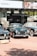 A selection of classic cars displayed outdoors on a brick platform, beneath a large Sotheby's auction sign. The scene includes a row of well-maintained vintage vehicles, including green and black models, with a background of promotional banners for a luxury online automotive auction.