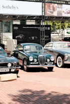 A selection of classic cars displayed outdoors on a brick platform, beneath a large Sotheby's auction sign. The scene includes a row of well-maintained vintage vehicles, including green and black models, with a background of promotional banners for a luxury online automotive auction.