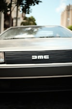 A classic car front with a distinctive grille featuring the DMC logo. The scene includes blurred surroundings with some urban elements like buildings and trees in the background.