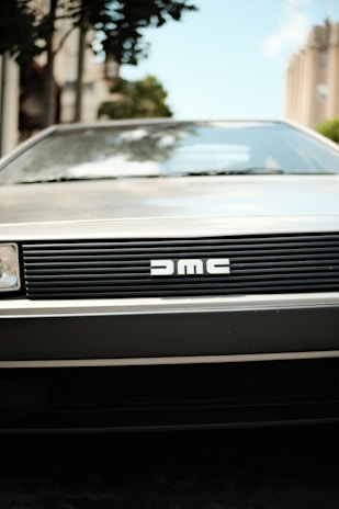 A classic car front with a distinctive grille featuring the DMC logo. The scene includes blurred surroundings with some urban elements like buildings and trees in the background.