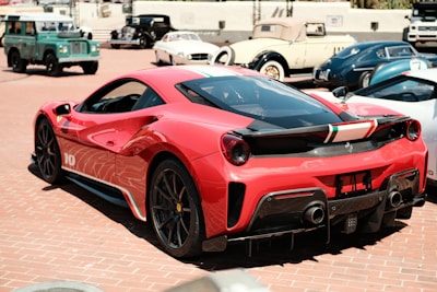 A sleek red sports car with aerodynamic curves and vivid details is parked prominently in the foreground. The car exhibits a racing stripe with the colors of the Italian flag. Surrounding it are several classic cars, including a green off-road vehicle and a vintage white convertible, set against a sunlit brick-paved area.