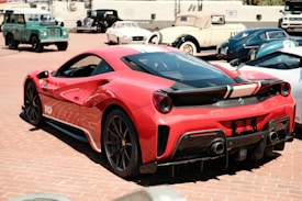 A sleek red sports car with aerodynamic curves and vivid details is parked prominently in the foreground. The car exhibits a racing stripe with the colors of the Italian flag. Surrounding it are several classic cars, including a green off-road vehicle and a vintage white convertible, set against a sunlit brick-paved area.