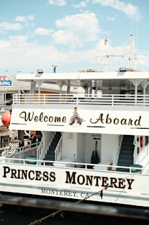 A large boat with signage reading 'Welcome Aboard' and 'Princess Monterey' is docked. The name 'Monterey, CA' is painted on the side, and there are stairs leading to an upper deck. An American flag and marine-related decorations are visible.