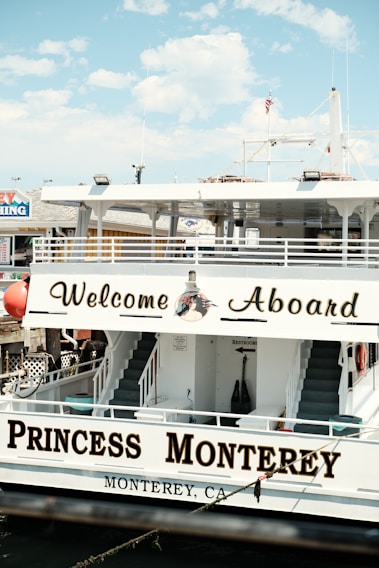 A large boat with signage reading 'Welcome Aboard' and 'Princess Monterey' is docked. The name 'Monterey, CA' is painted on the side, and there are stairs leading to an upper deck. An American flag and marine-related decorations are visible.