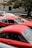 Several vintage sports cars are parked under the shade of a large tree beside a building. The cars have a classic design, with vibrant red colors dominating the scene. There's a mix of coupe and convertible models, showcasing their distinct curves and body styles.